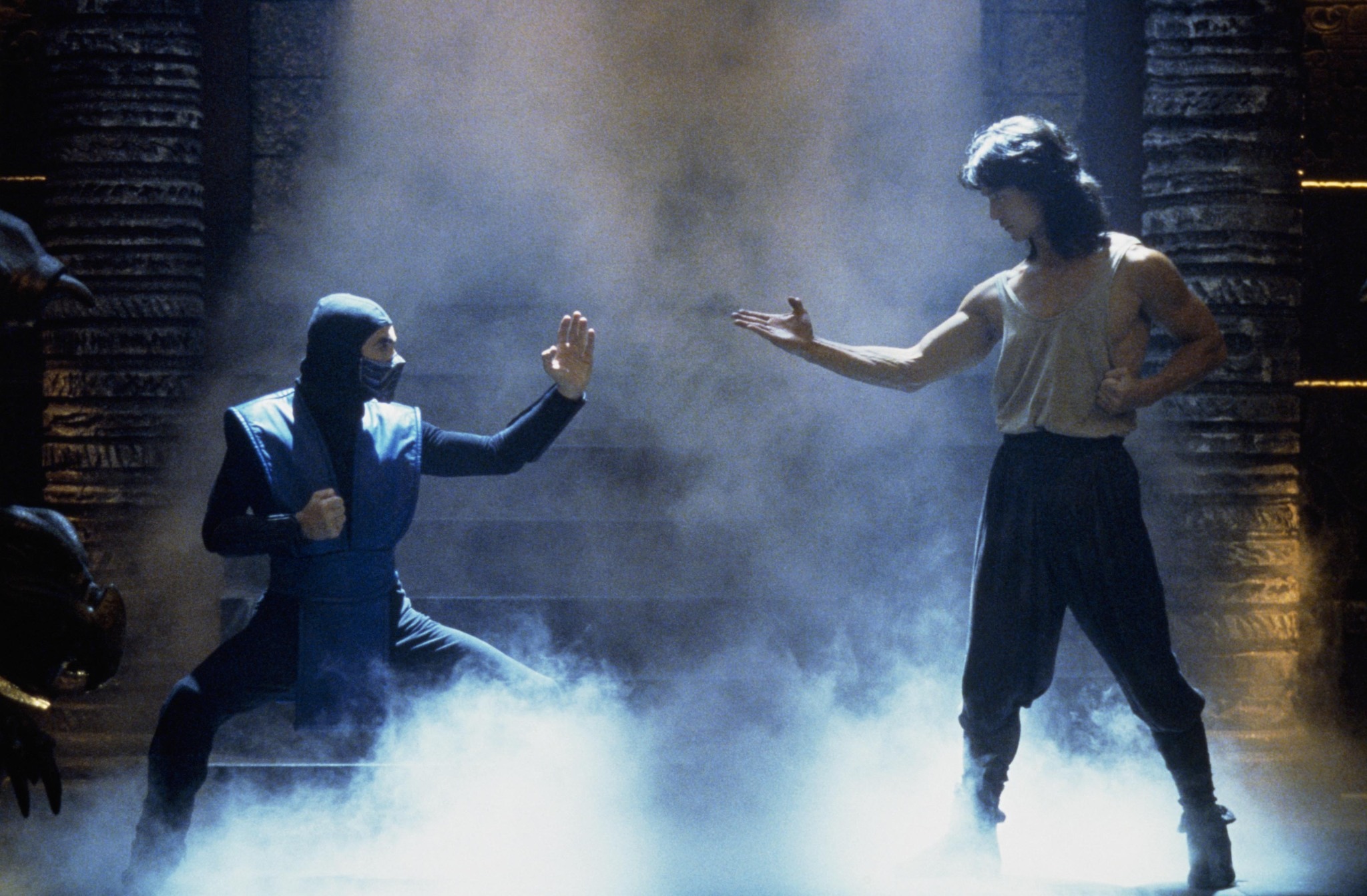 Sub-Zero, dressed in his blue ninja outfit, in a fighting stance against Liu Kang played by Robin Shou, who is in a martial arts pose with a misty, dimly lit background.