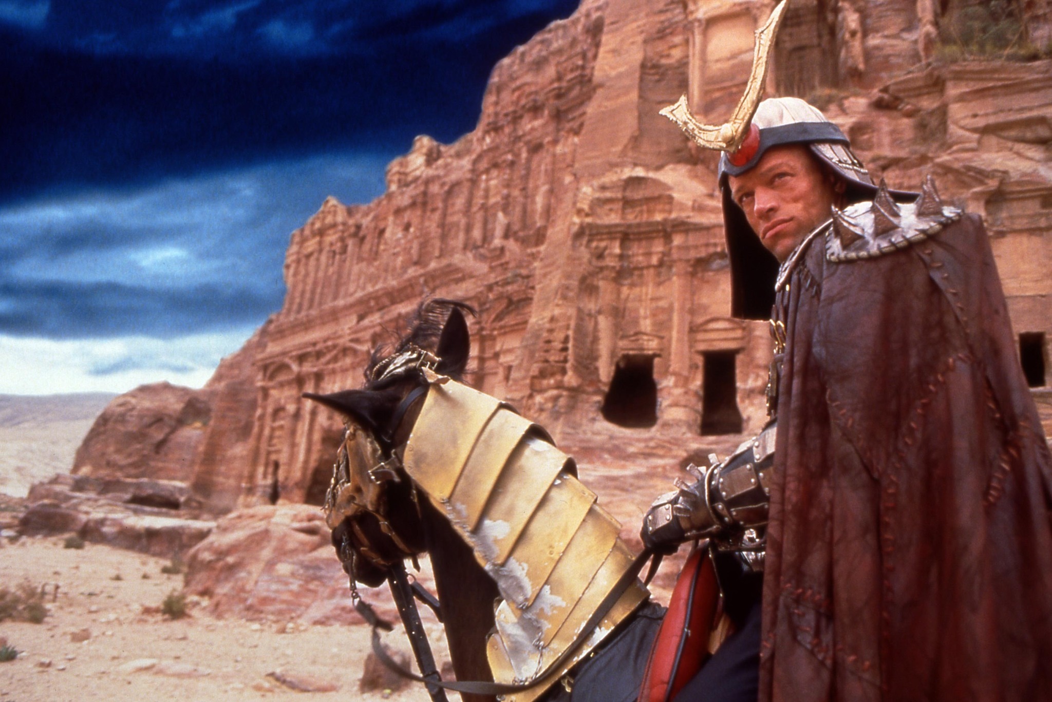 Warlord Shao Kahn in a spiked warrior helmet and cloak, riding an armored horse against a desert landscape with ancient ruins in the background.