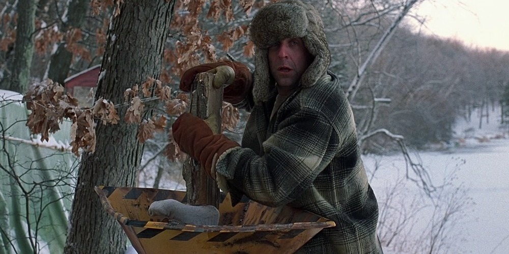 Peter Stormare as Gaear Grimsrud wearing a fur hat and winter coat while disposing of a body using a woodchipper in a chilling and infamous scene from the Coen brothers’ crime thriller.